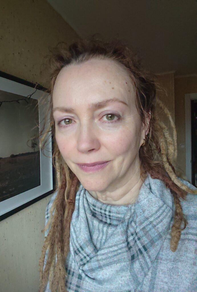 A white woman with brown eyes and blonde dreadlocks, wearing a grey dress